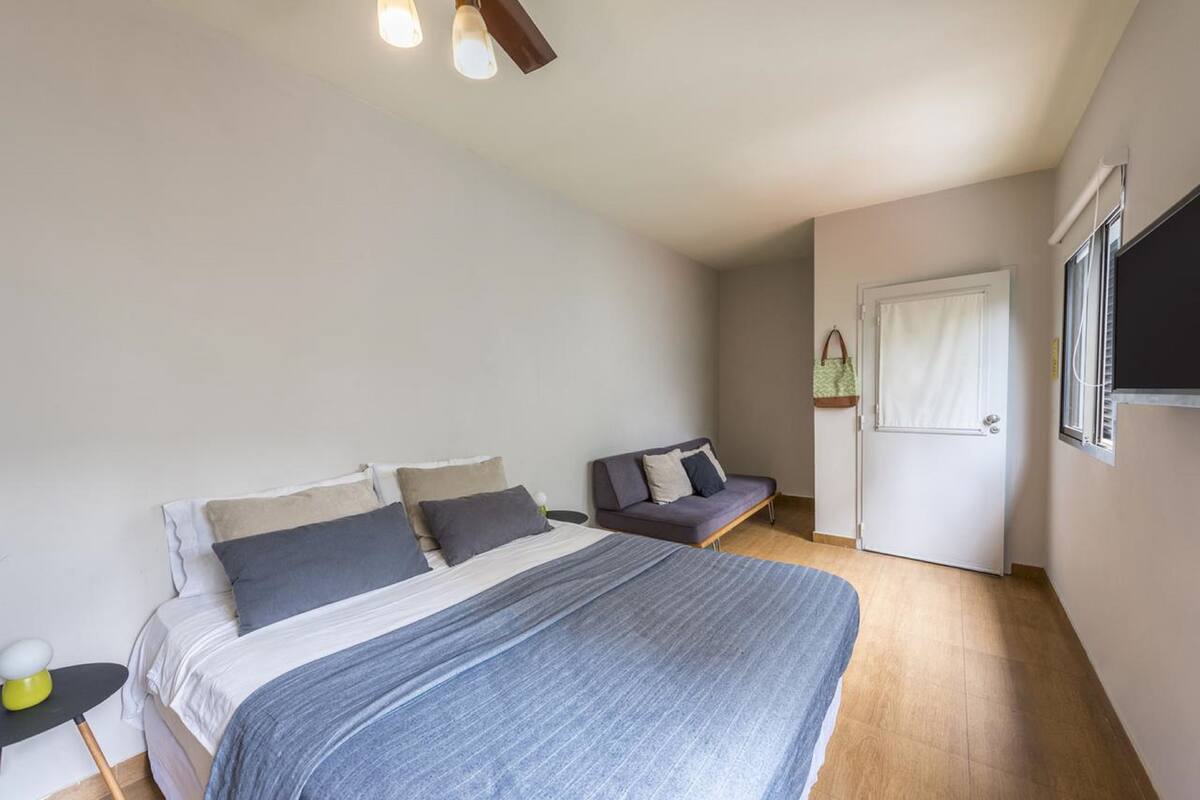 A bedroom with a large bed, a smaller sofa, a bedside table, a door, and a window.