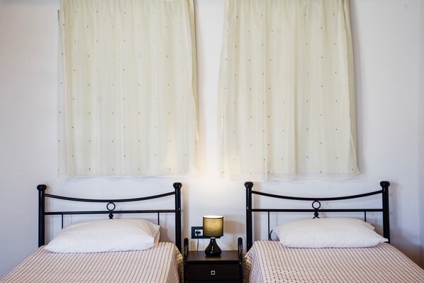 A double bed with a nightstand and a lamp, two white pillows, and sheer curtains on a white wall. - Estrella Villas (Rhodes)