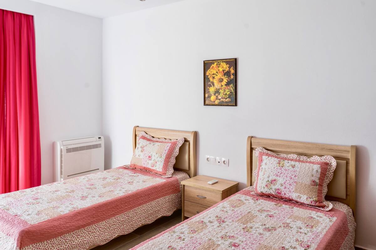 Un dormitorio con dos camas, una cortina roja, un aire acondicionado instalado en la pared, una mesita de noche con una lámpara y una obra de arte floral enmarcada en la pared.