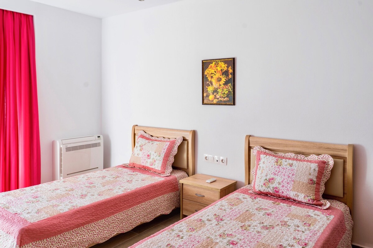 Ein Schlafzimmer mit zwei Betten, einem roten Vorhang, einer Wand-Klimaanlage, einem Nachttisch mit Lampe und einem gerahmten Blumenbild an der Wand.