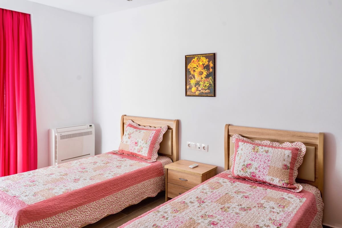 Una habitación con dos camas, una cortina roja, aire acondicionado instalado en la pared, una mesita de noche con un control remoto y un cuadro enmarcado en la pared.