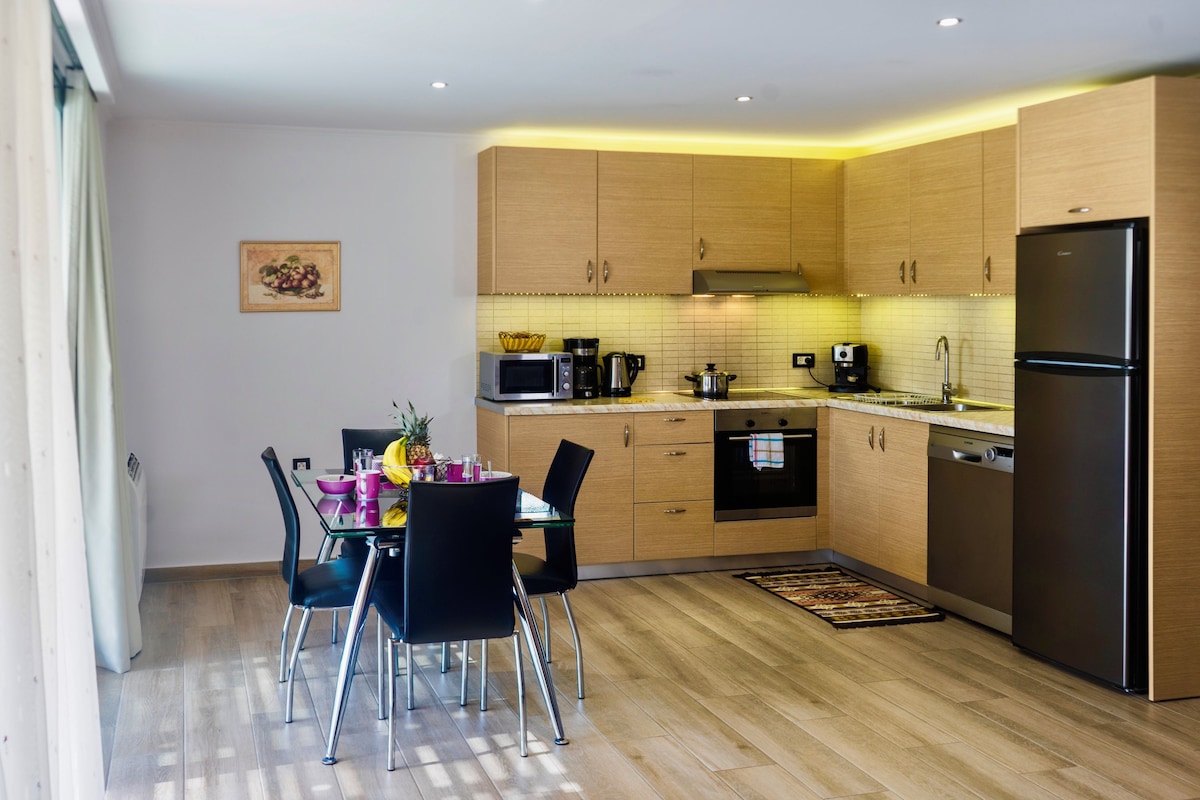 Uma cozinha moderna com armários de madeira, eletrodomésticos de aço inoxidável e uma área de refeições com cadeiras pretas e uma mesa de vidro.