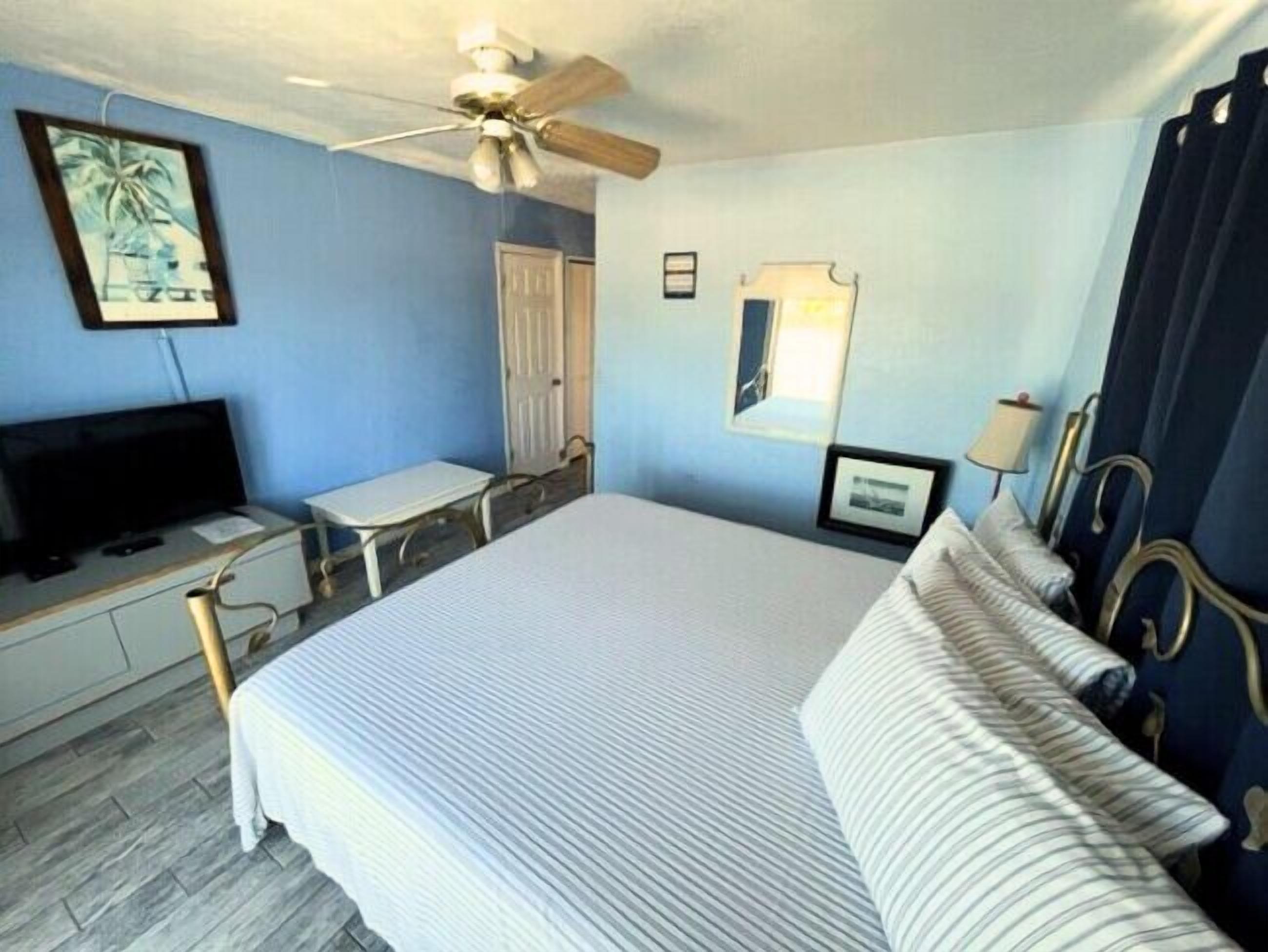 A bedroom with a bed, a desk, a television, a ceiling fan, and a window with curtains.