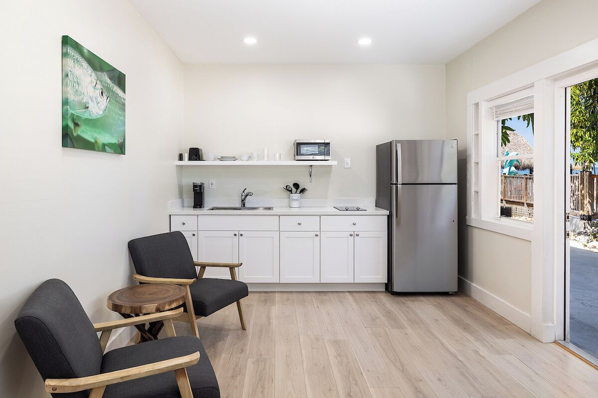 A compact kitchen with white cabinets, a stainless steel refrigerator, a microwave, and a sink. There are two dark chairs and a small round table. A painting of a fish is on the wall.