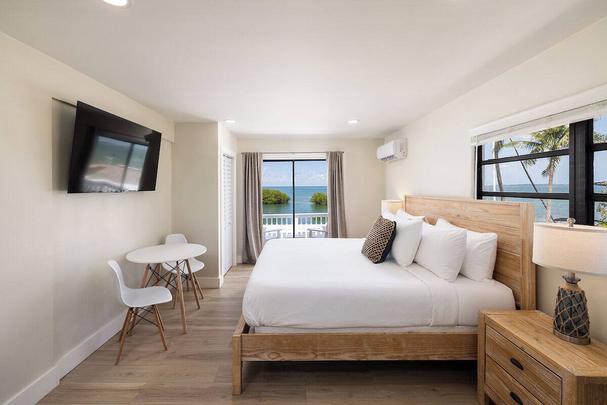 A hotel room with a bed, a TV, a balcony with a view of the sea, and a table with chairs.