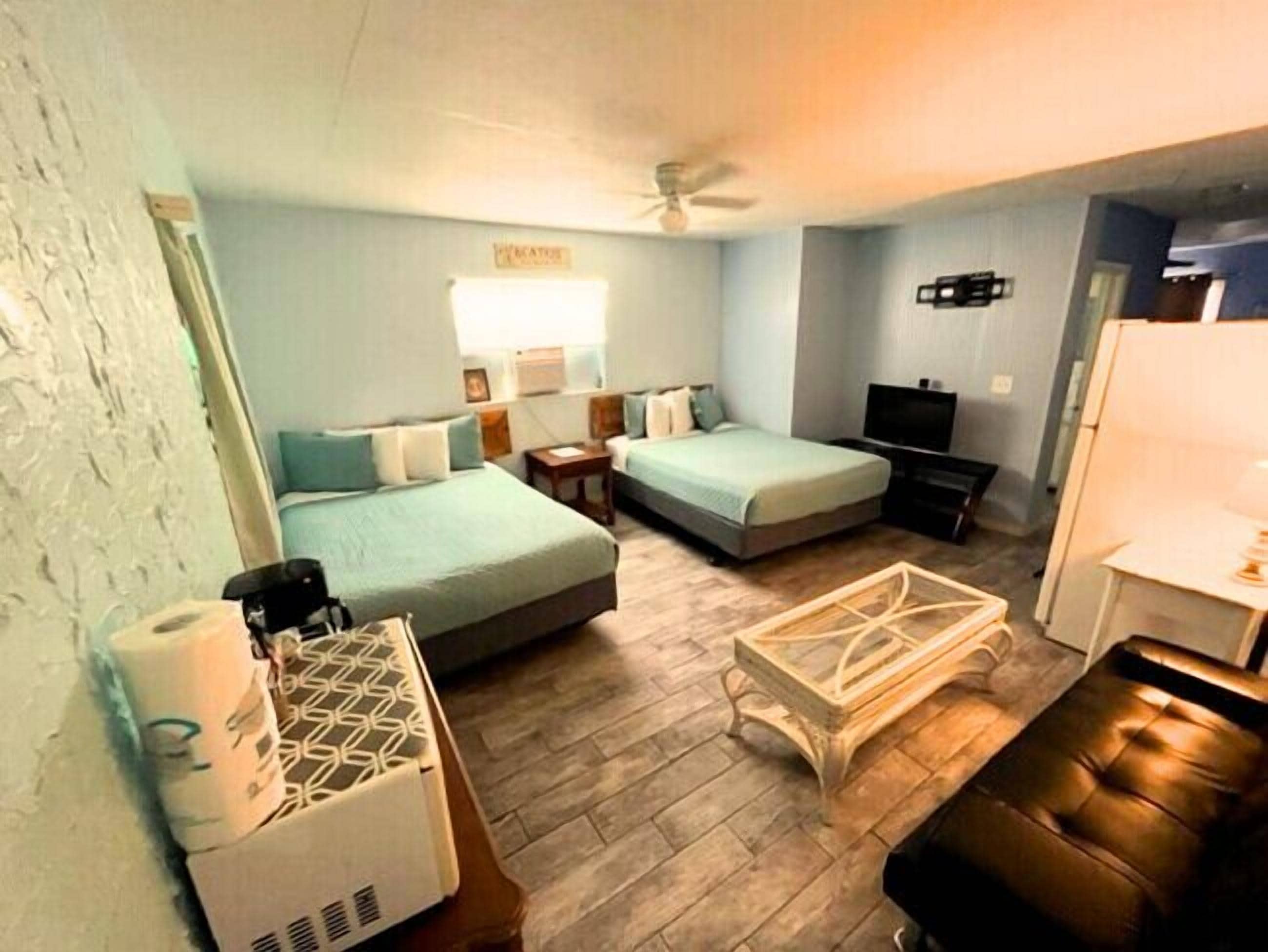 A compact hotel room with two beds, a TV, a ceiling fan, and a kitchenette.