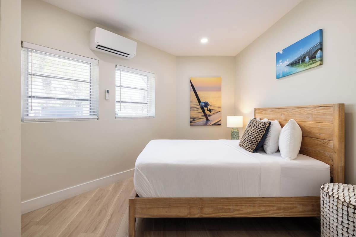 A bedroom with a wooden bed, two windows with blinds, an air conditioner, a painting of a sunset, and a lamp.