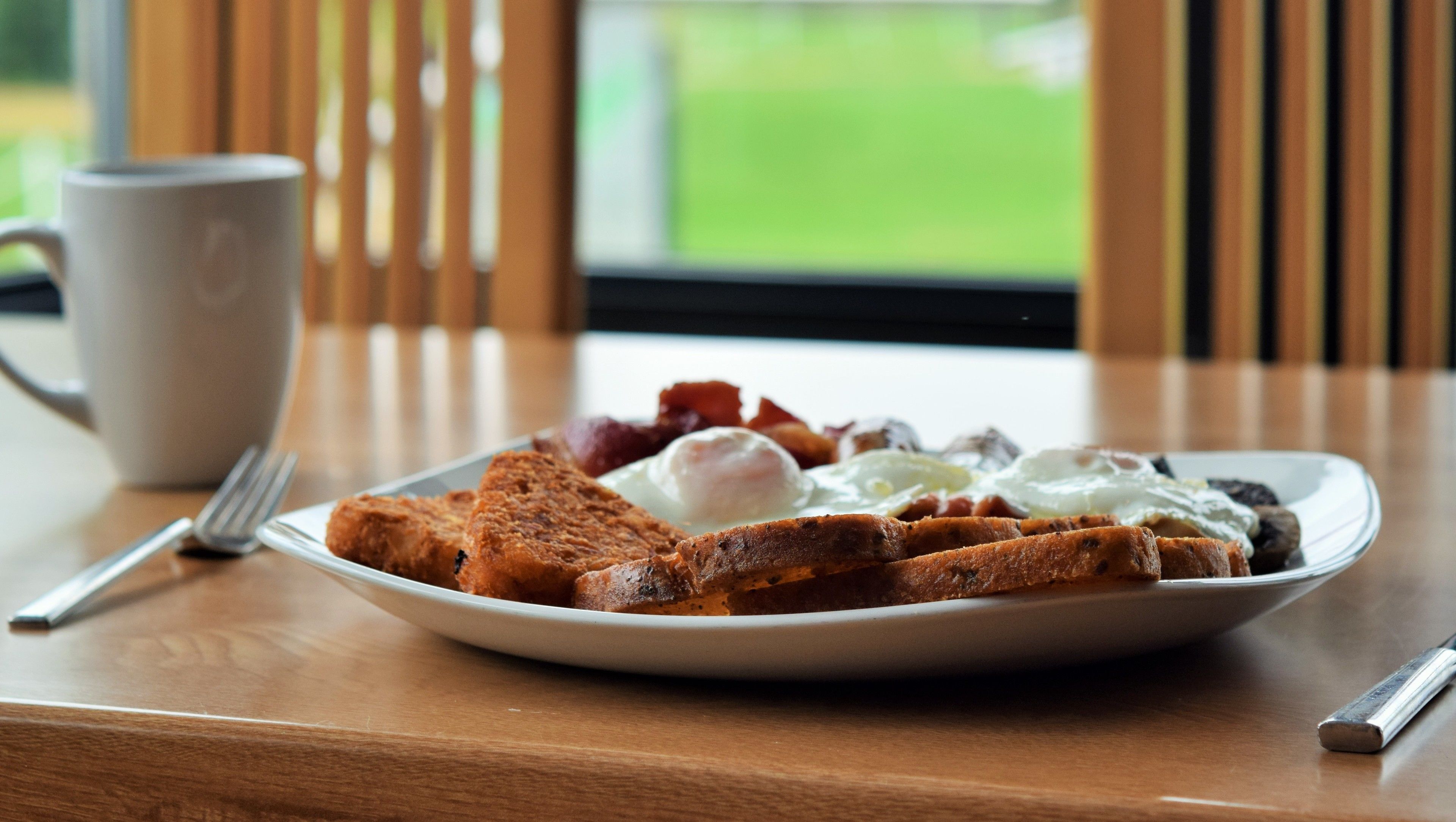 A breakfast plate with toast, eggs, and bacon on a wooden table with a white mug and cutlery.
