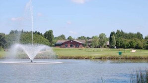Un lac paisible avec une fontaine, entouré d’une végétation luxuriante et bordé d’un bâtiment à toit rouge.