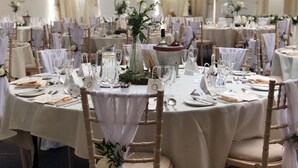 Une salle de banquet avec des tables rondes dressées pour un événement formel, chacune agrémentée de marque-places, de verres et de compositions florales.