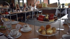 Un présentoir à trois niveaux garni de diverses pâtisseries et de sandwichs, une table avec des tasses et une théière, et un groupe de personnes à l’arrière-plan.