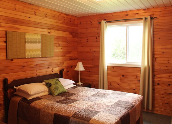 Habitación de cabaña de madera con cama, mesita de noche, lámpara y ventana con cortinas.