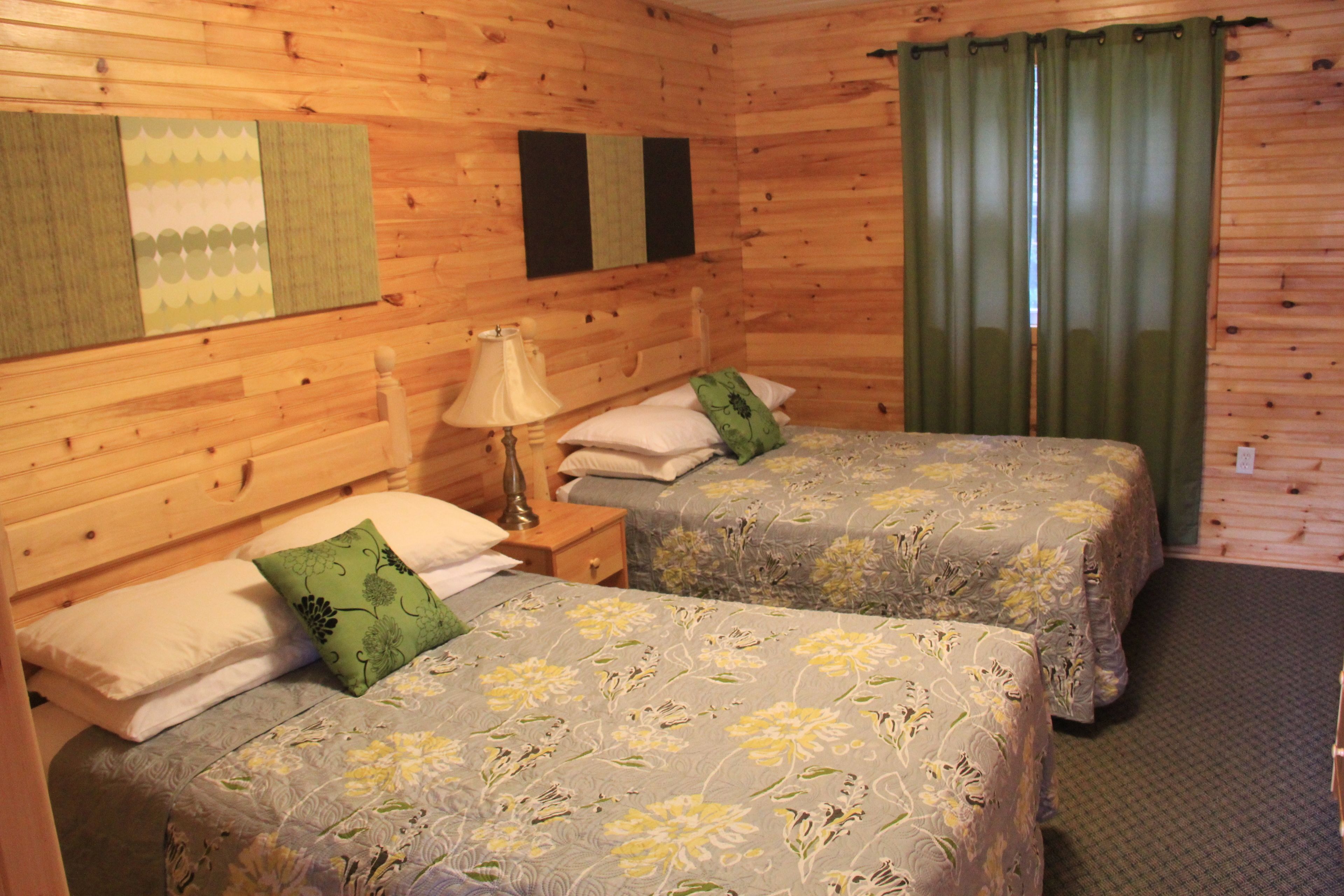 A wooden cabin room with two beds, each with floral bedding, a nightstand with a lamp, and a green curtain.