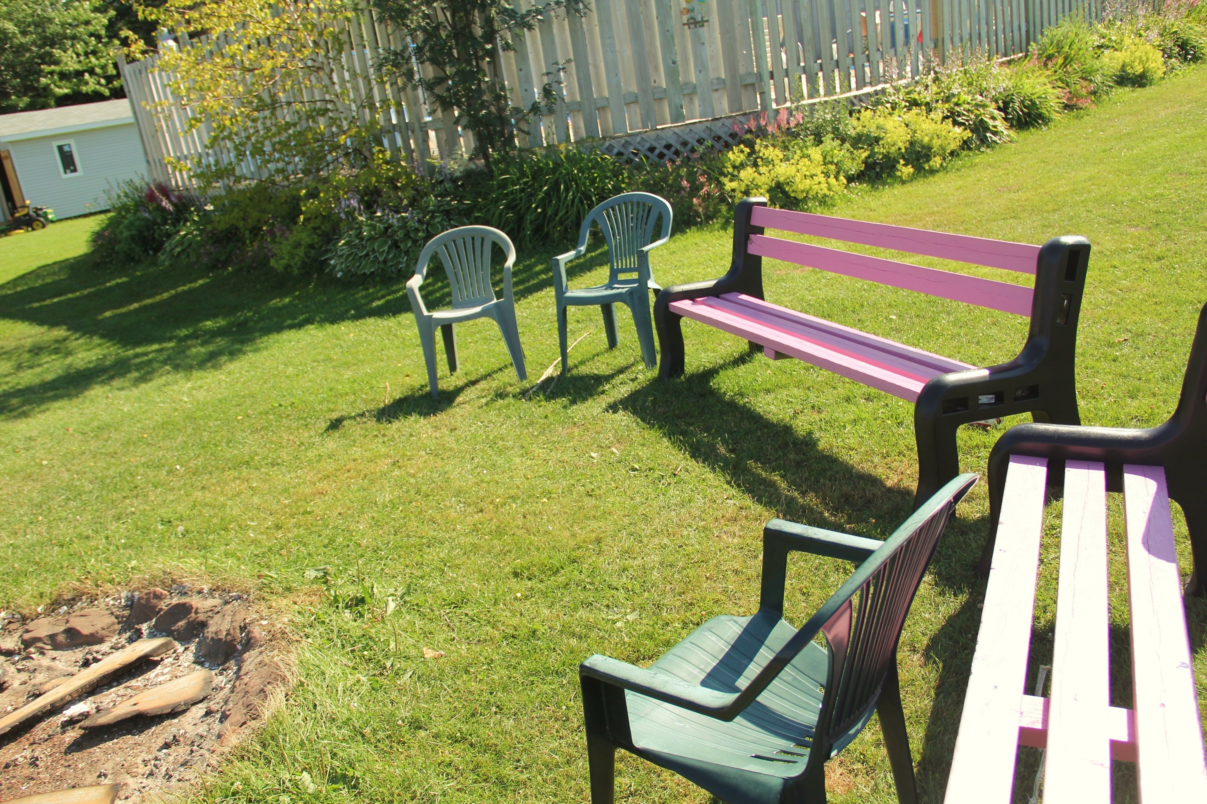 A grassy outdoor area with a wooden bench and two plastic chairs.