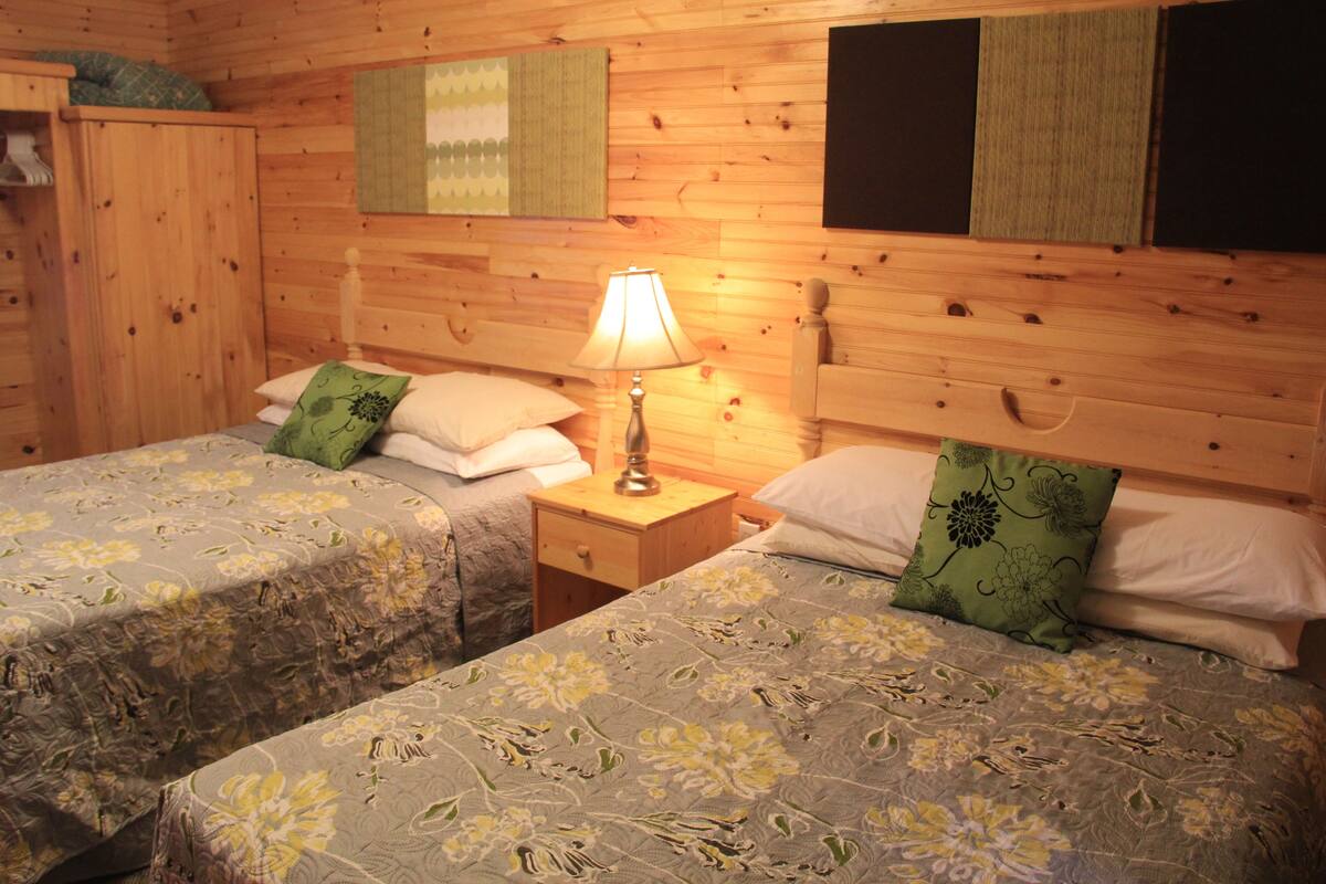 Ein Zimmer mit zwei Betten, jeweils mit Blumenmuster bezogen und grünen Kissen. Ein Holzschrank und ein Nachttisch mit Lampe sind ebenfalls vorhanden.