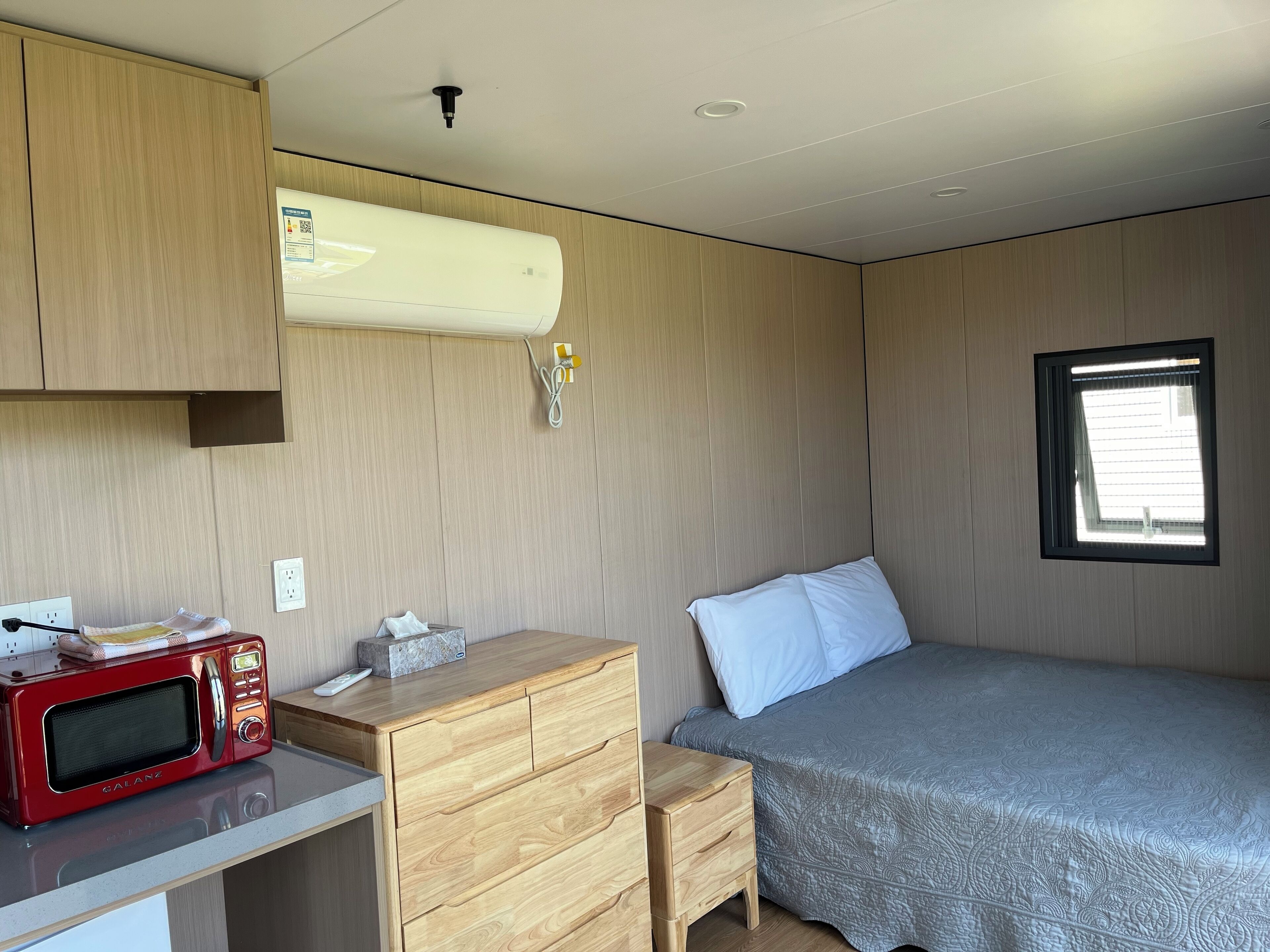 A compact room with a bed, a wooden dresser, a red microwave, and a window.