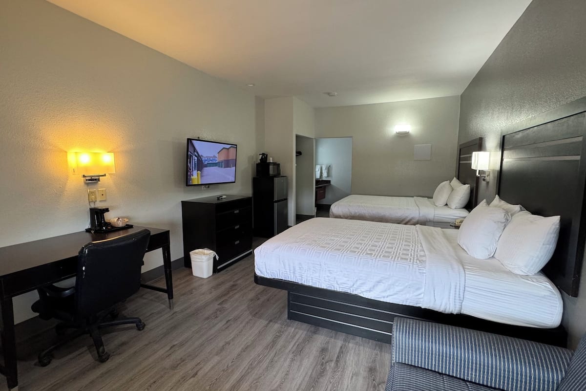 A hotel room with two beds, a desk, a chair, a television, and a small kitchenette.