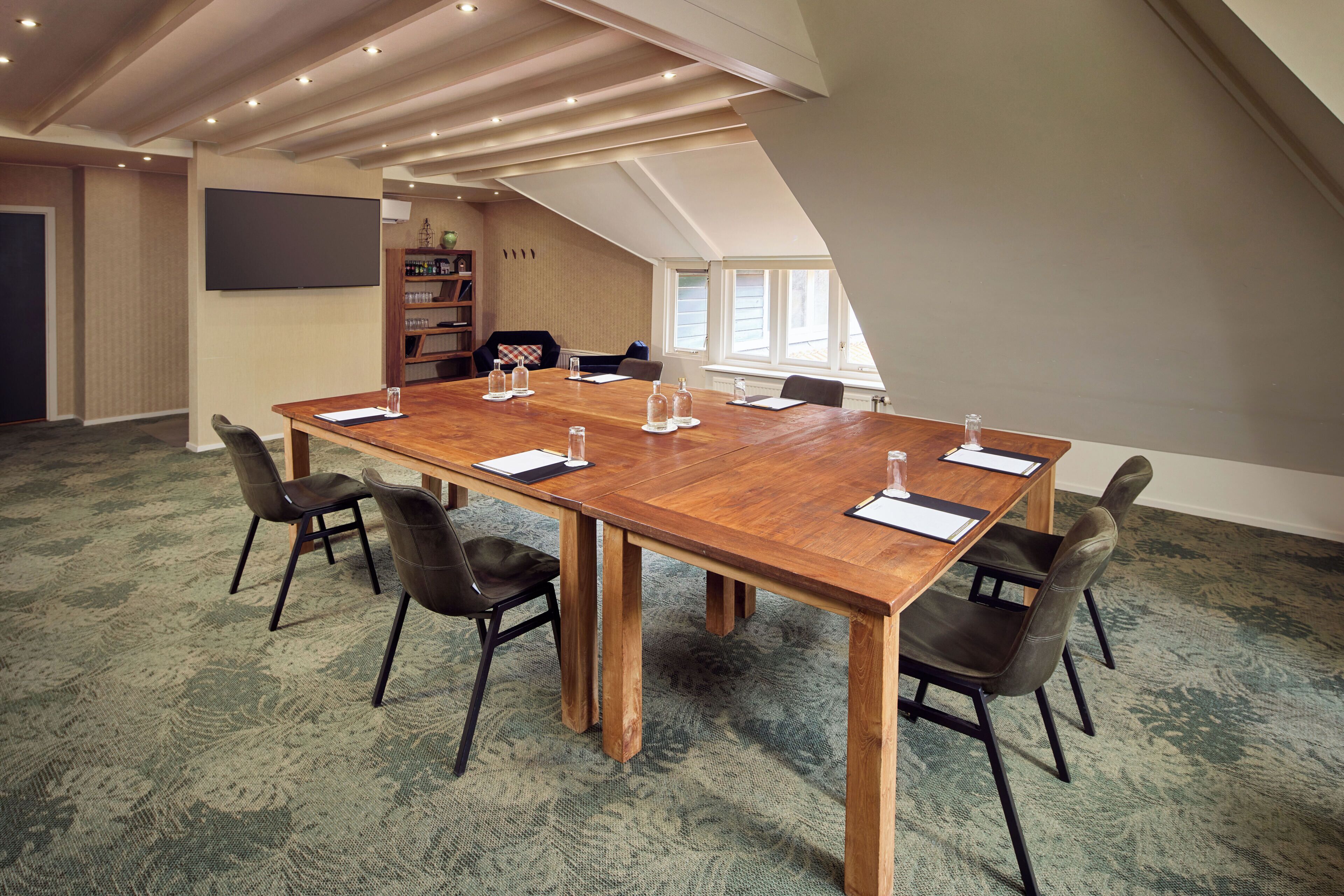 A conference room with a large wooden table, six chairs, a bookshelf, and a large window.