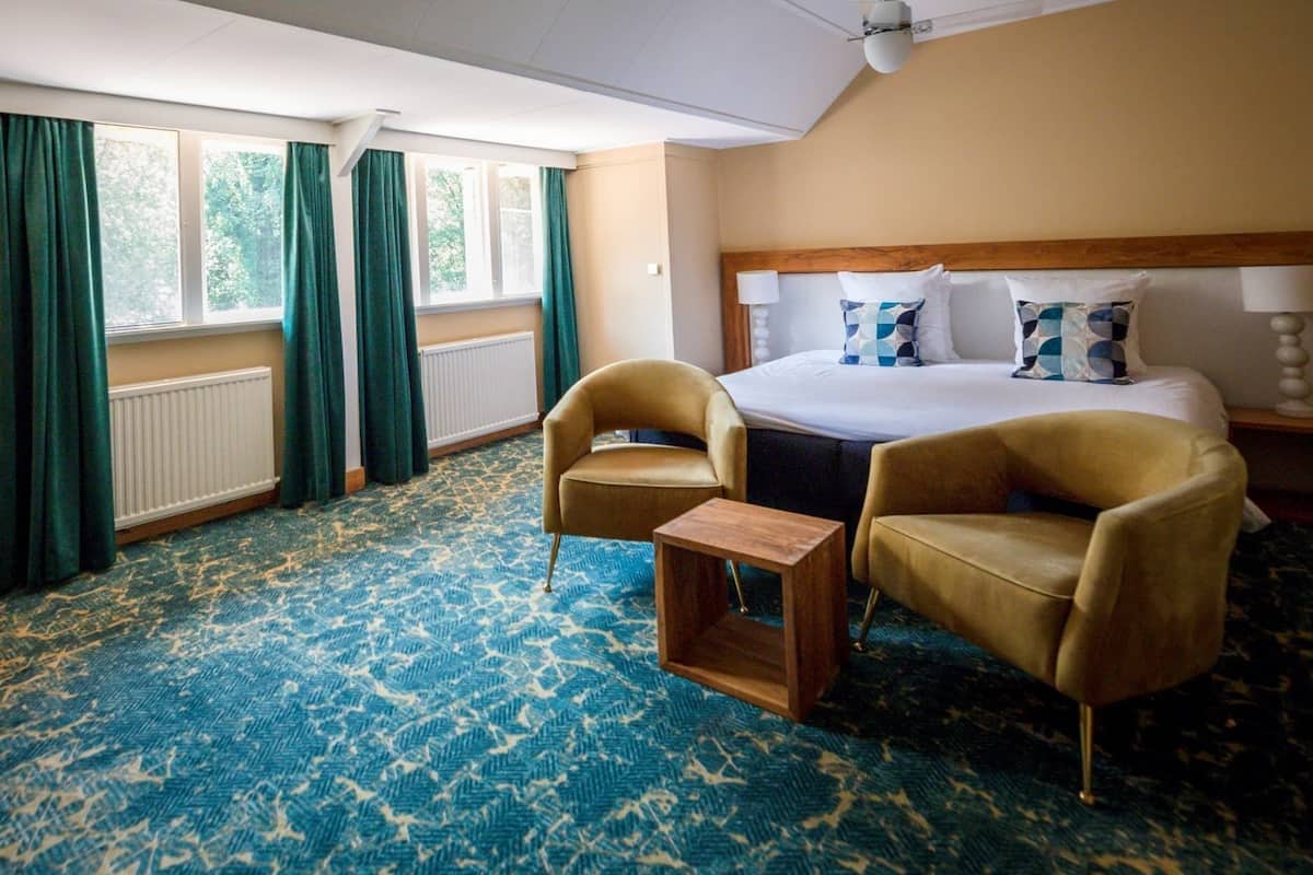 A hotel room with a bed, two armchairs, a small table, a radiator, and a window with curtains.