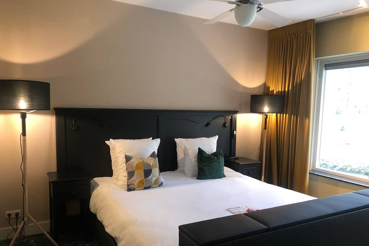 A hotel room with a large bed, two bedside tables, a ceiling fan, and a window with curtains.