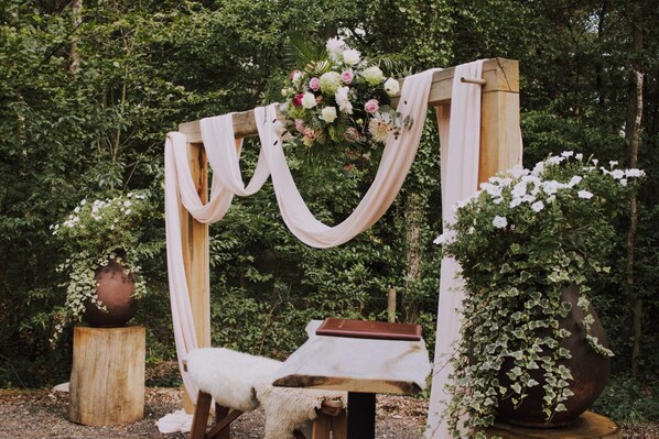 A wooden archway with draped fabric and floral arrangements, a bench with a book, and potted plants.