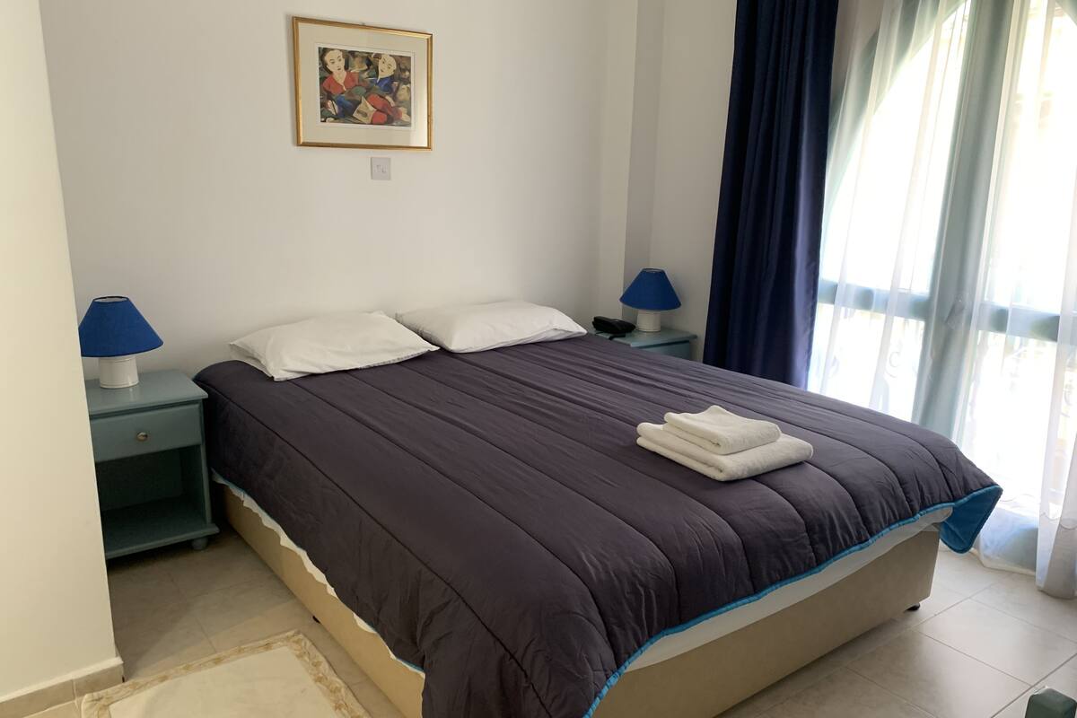 Un lit bien fait, recouvert d’une couette de couleur sombre, avec deux oreillers blancs et une serviette blanche pliée posée sur le dessus. Deux tables de chevet avec des lampes se trouvent de chaque côté du lit. Un tableau encadré est accroché au mur, au-dessus du lit.