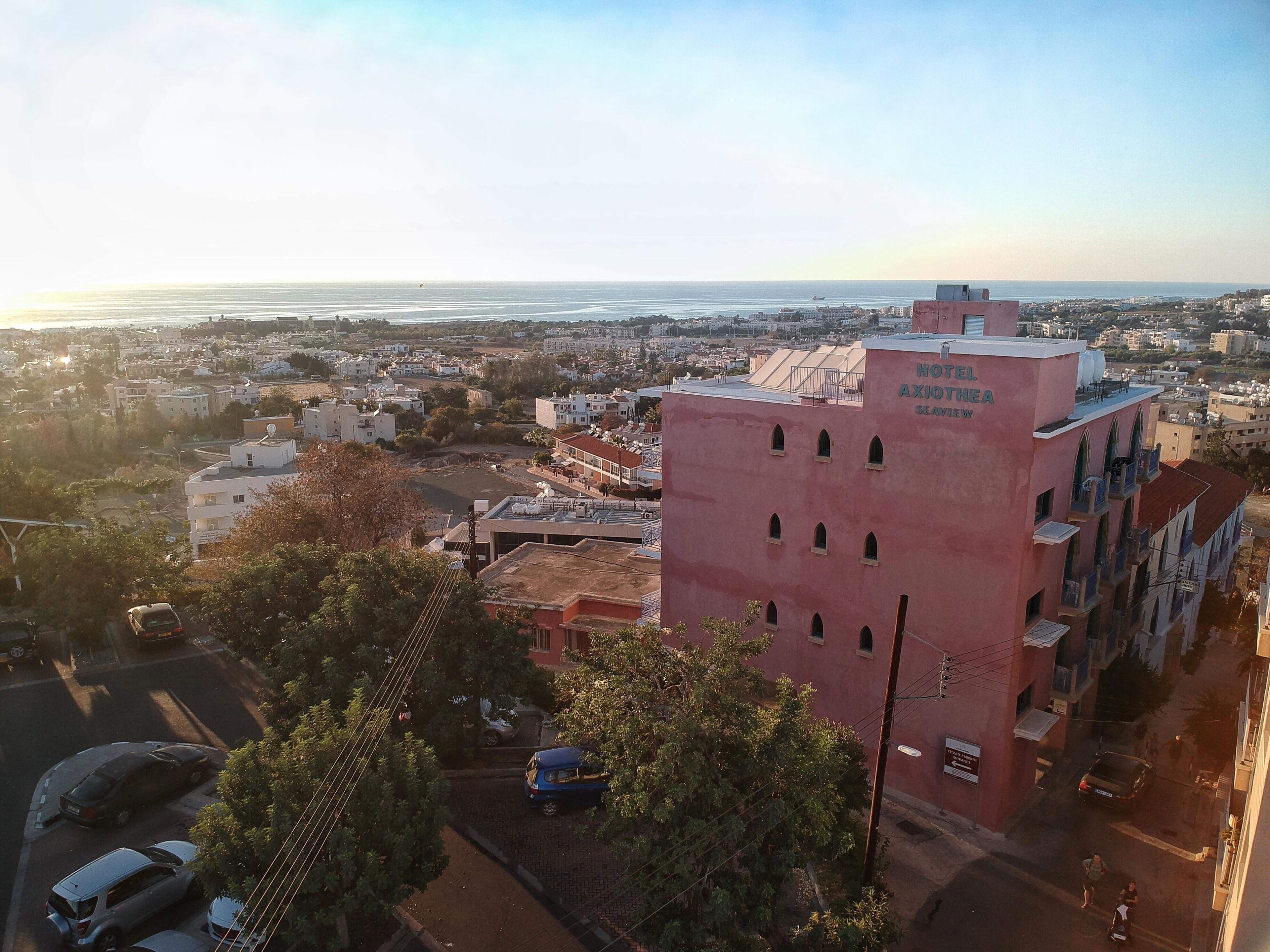 A pink building with the name "HOTEL AXIOTHEA" and a view of a coastal cityscape.