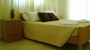 A single bed with a quilted cover, two pillows, a wooden nightstand with drawers, and a bedside table. - Aurora Apartments (Pegeia)