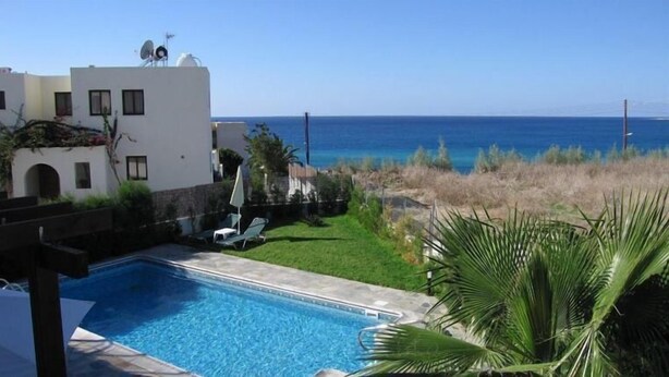 A swimming pool with lounge chairs, a view of the sea, and a building with satellite dishes. - Aurora Apartments (Pegeia)