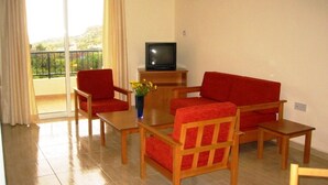 A living room with a sofa, two armchairs, a coffee table, and a TV on a wooden stand. - Aurora Apartments (Pegeia)