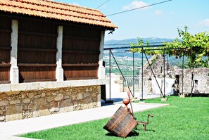 Un edificio con techo de tejas y paredes de piedra, un área verde con una estructura metálica y una vista de un paisaje montañoso al fondo.