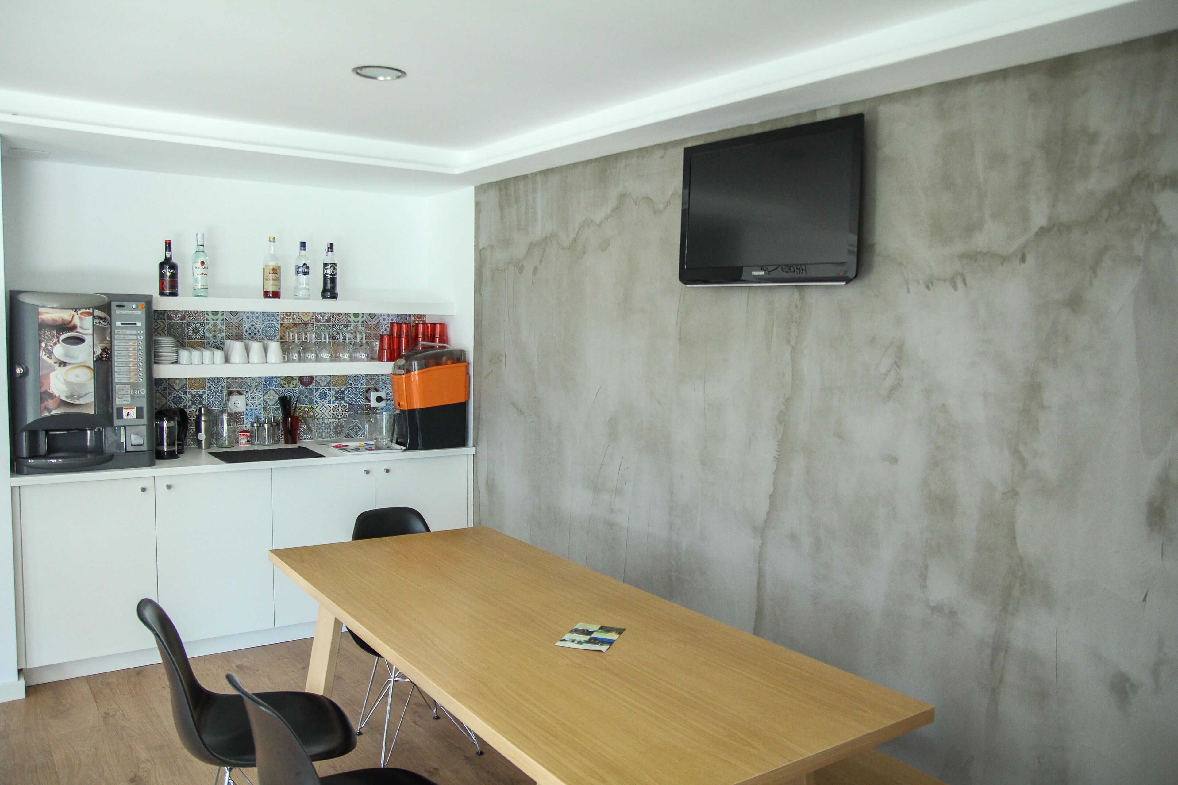 A modern kitchen with white cabinets, a wooden table, and a flat-screen TV mounted on a textured wall.