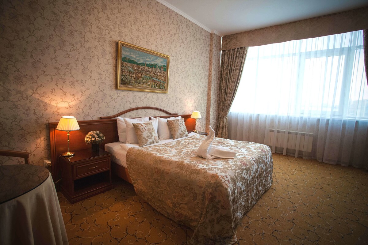 Une chambre d’hôtel avec un grand lit, deux lampes de chevet, une table de chevet avec une lampe, un tableau au mur et une fenêtre avec des rideaux.