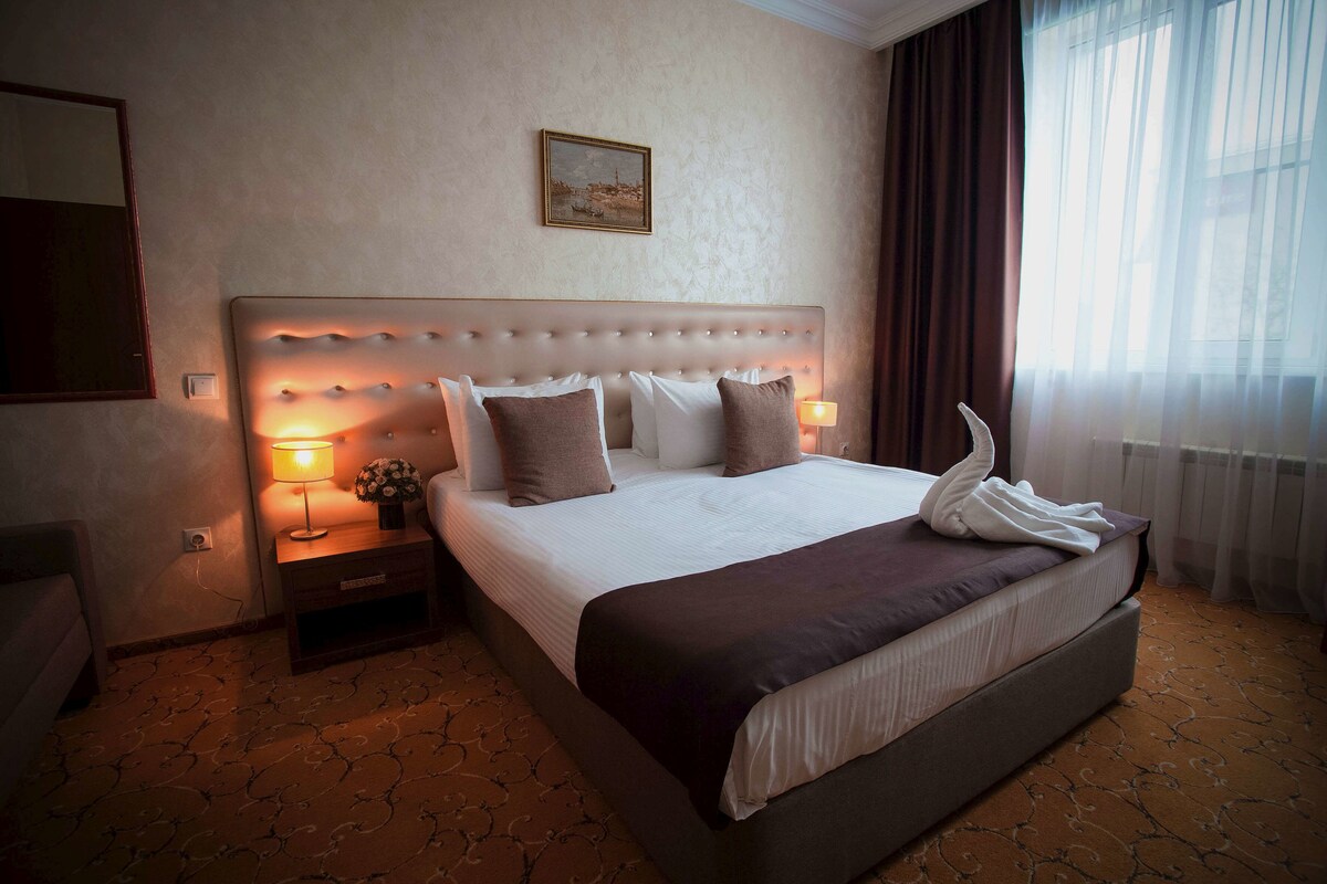 Une chambre d’hôtel avec un grand lit, une table de chevet avec une lampe, un tableau encadré au mur et une fenêtre avec des rideaux.
