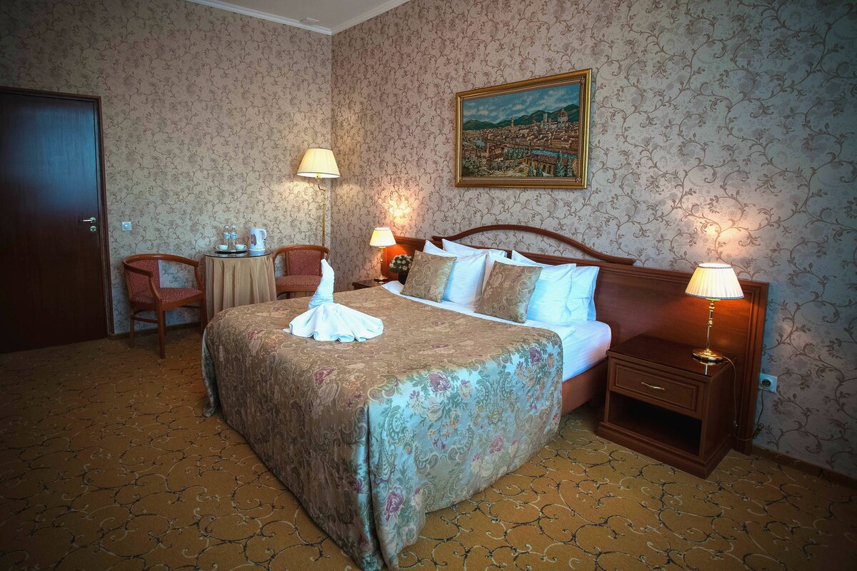 Une chambre d’hôtel avec un grand lit, deux tables de chevet avec des lampes, une chaise, une petite table avec une lampe et un tableau au mur.