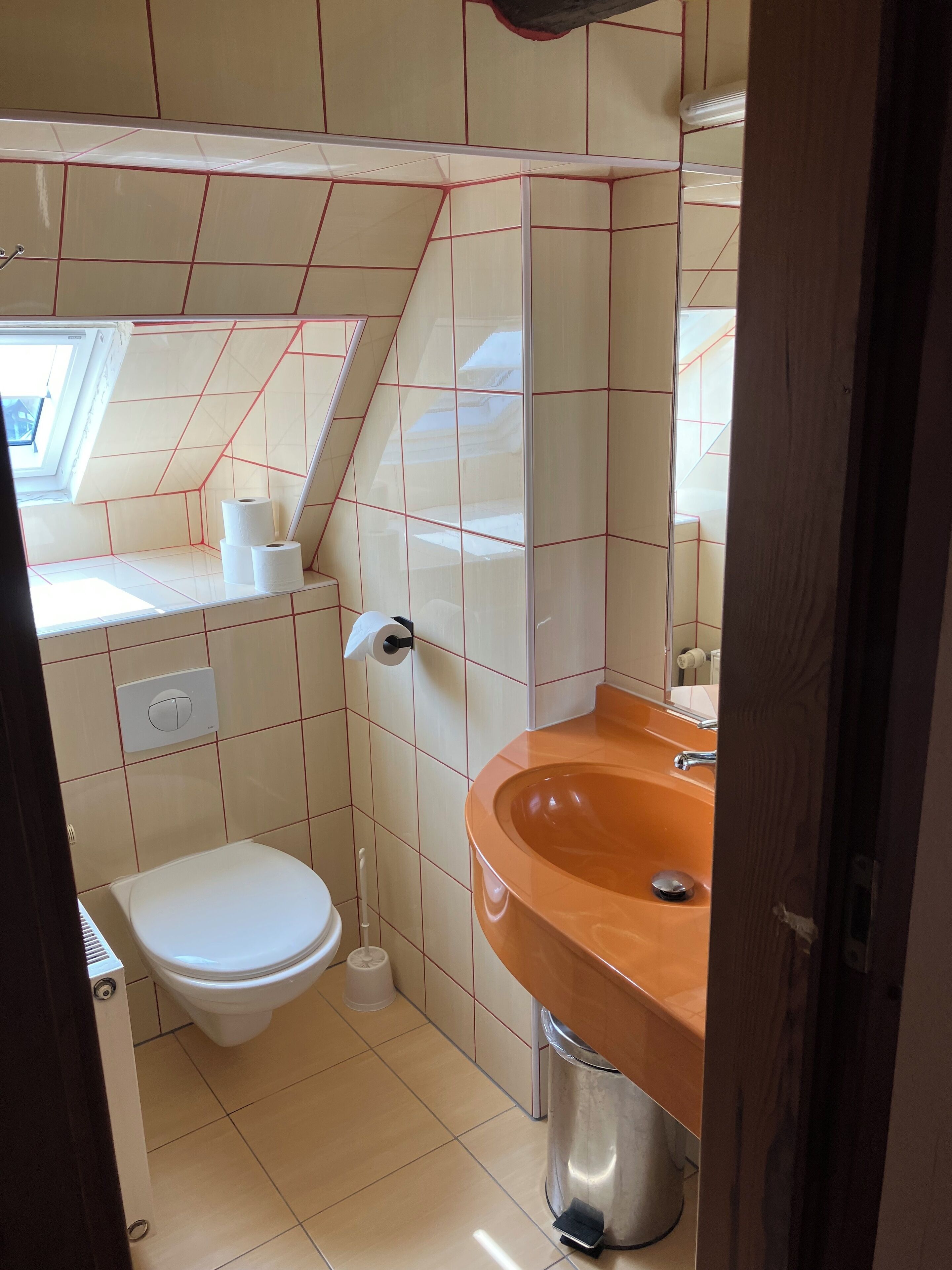 Et lille badeværelse med toilet, håndvask med brun skål og et vindue over toilettet.