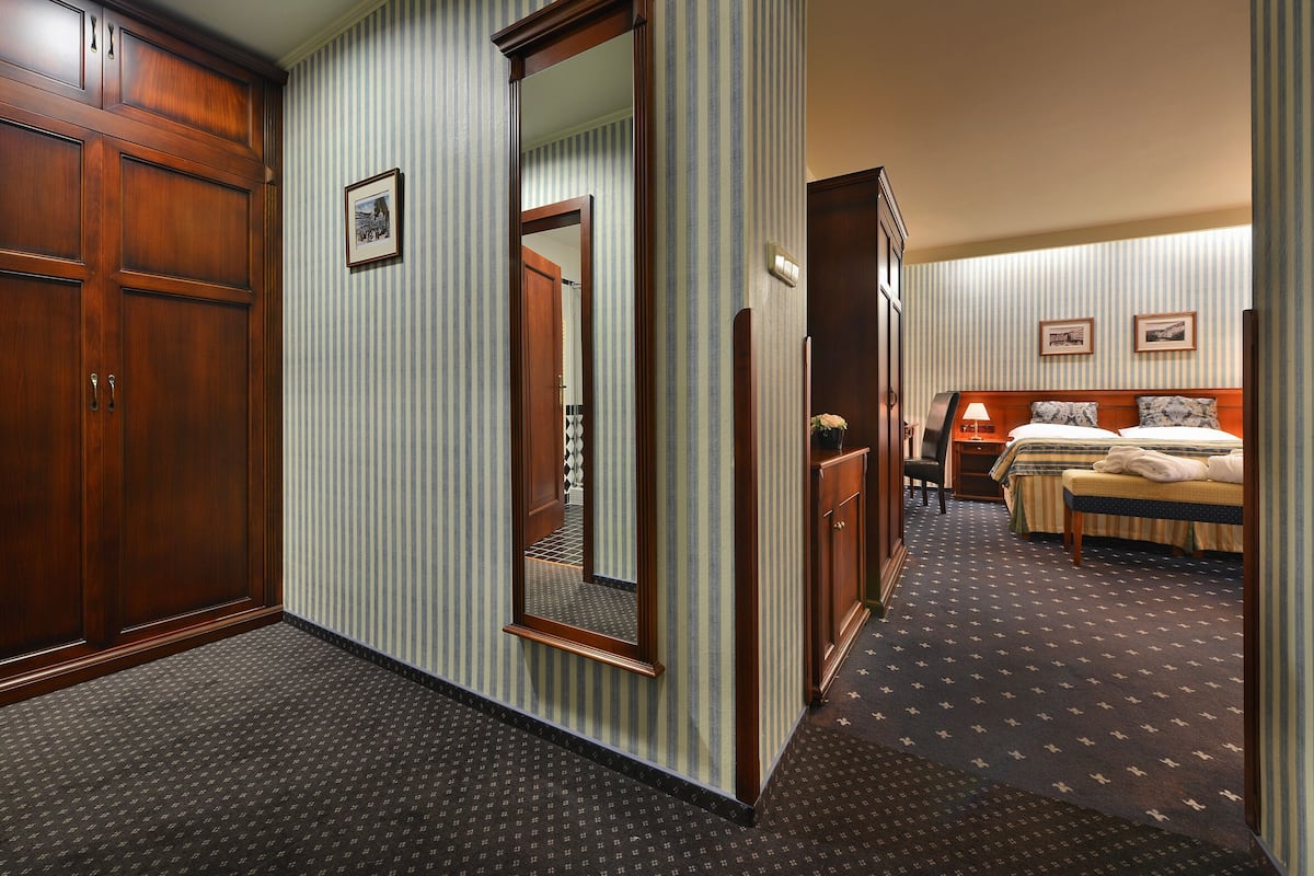 A hotel room with a large wooden wardrobe, a mirror, a bed with two pillows, a bedside table with a lamp, and a carpeted floor.