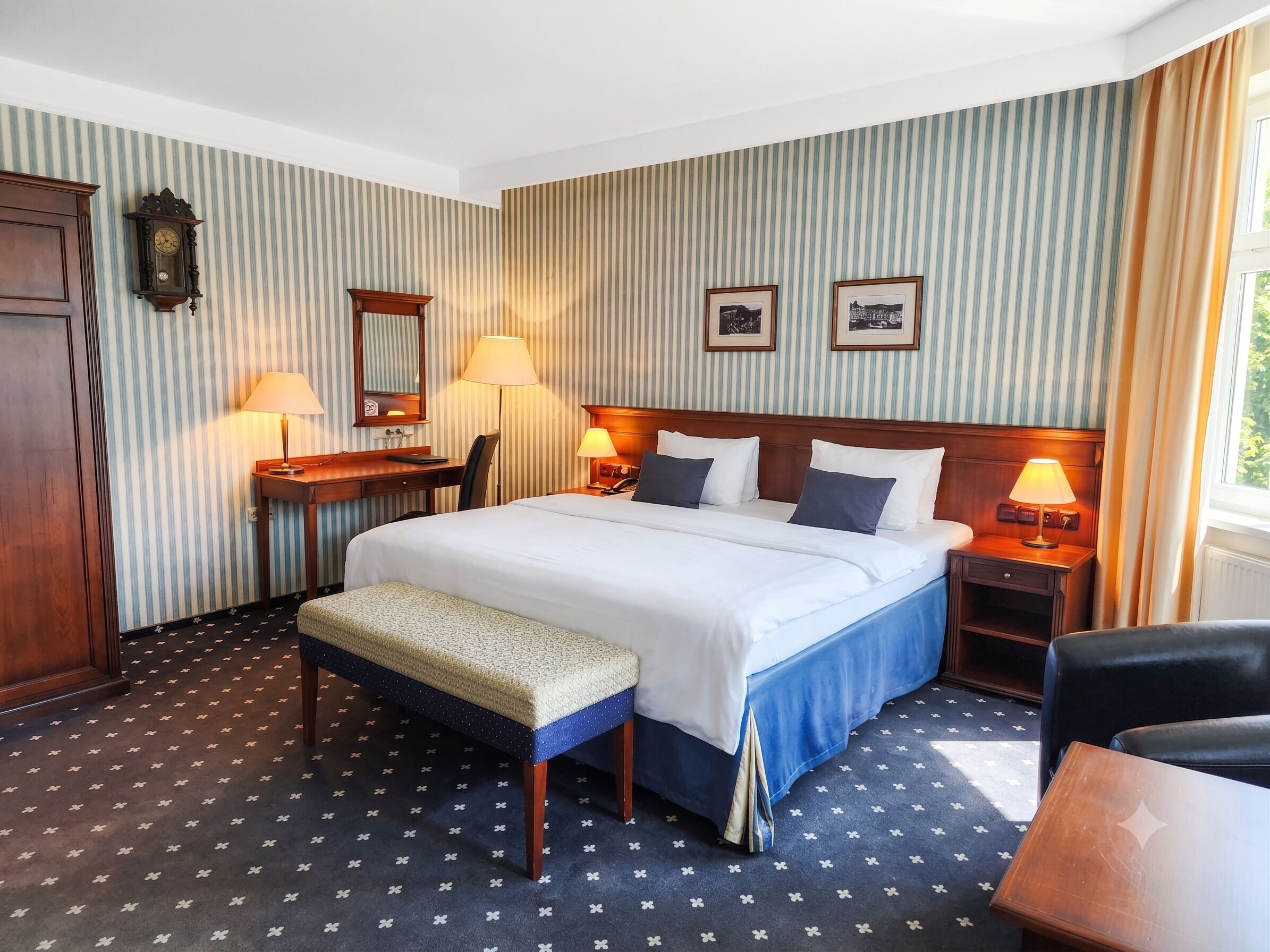 Habitación de hotel con una cama grande, mesitas de noche, un escritorio, una silla, un banco y un reloj en la pared.