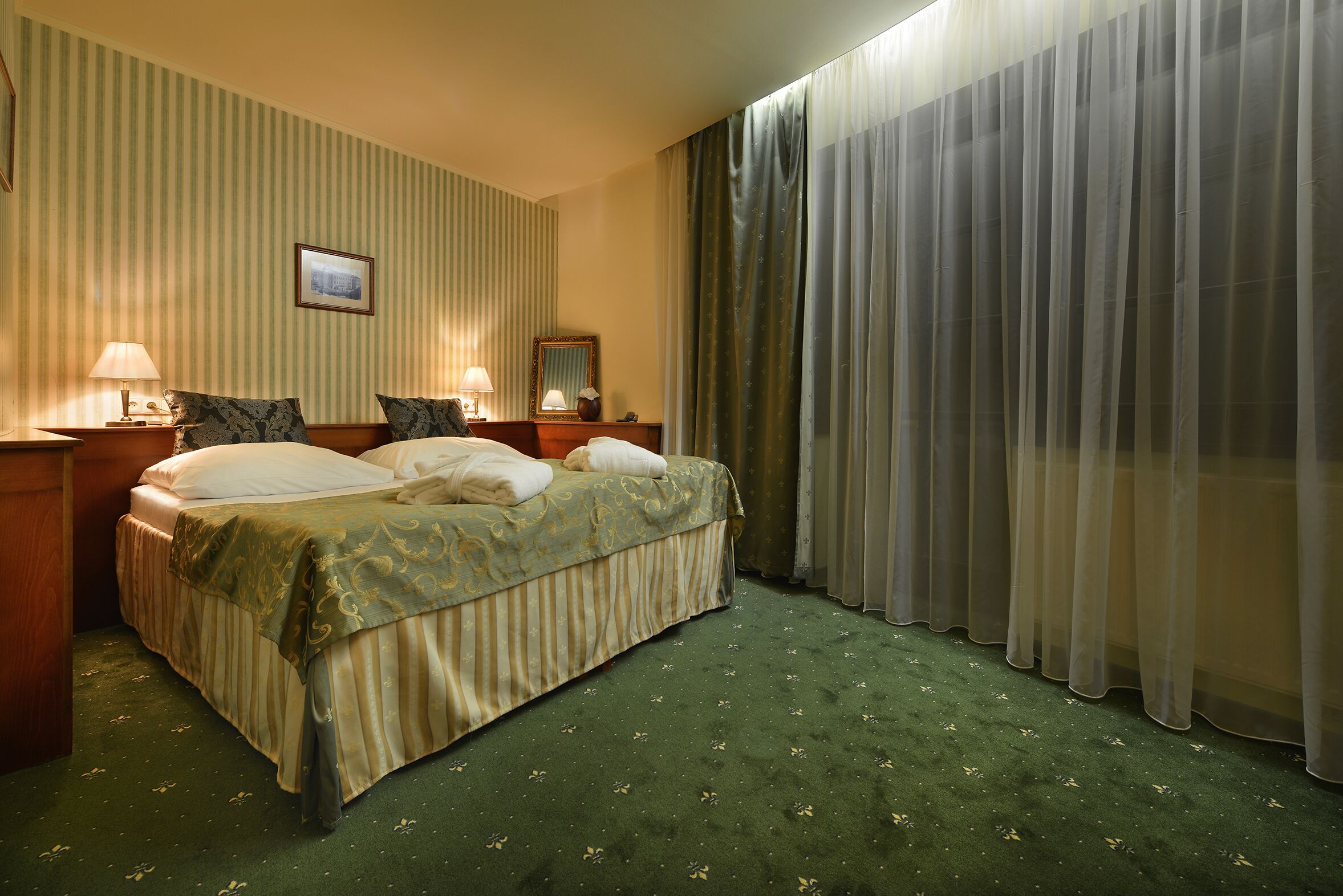 A hotel room with a bed, two bedside tables with lamps, a mirror, and a window with curtains.