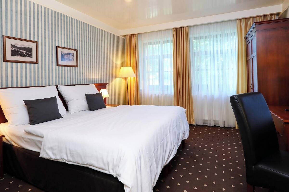 A hotel room with a large bed, two bedside tables with lamps, a desk with a chair, and two framed pictures on the wall.