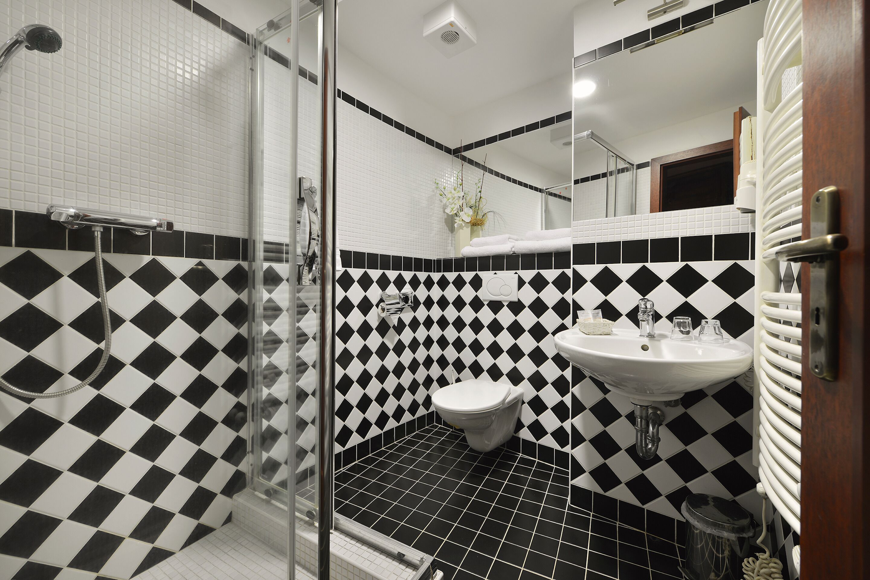 Un baño con suelo a cuadros blanco y negro, cabina de ducha de vidrio, lavabo blanco y retrete.