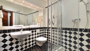 Un baño con piso a cuadros blancos y negros, un lavamanos blanco, un inodoro y una zona de ducha con mampara de vidrio.