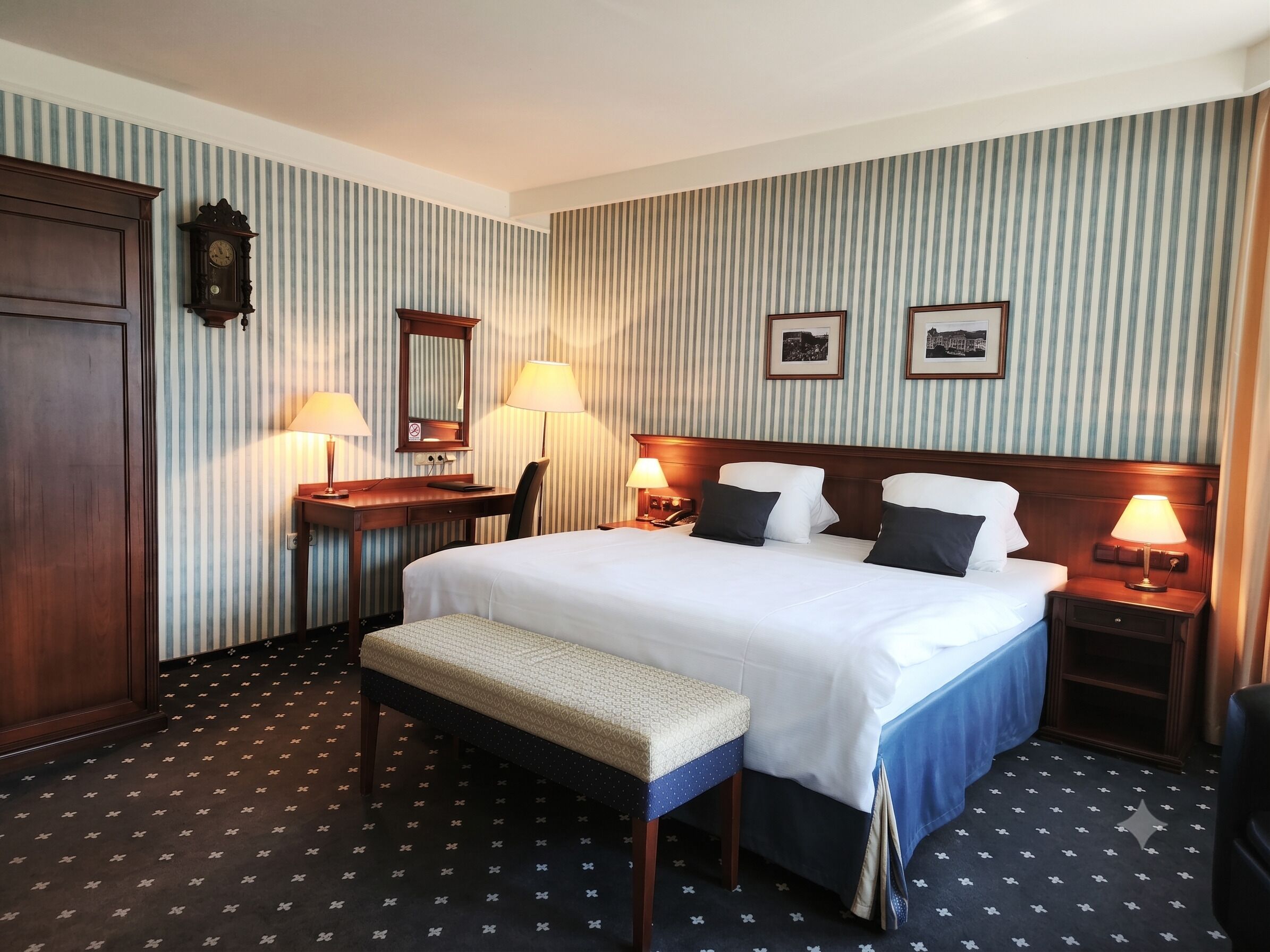 Habitación de hotel con una cama grande, mesitas de noche, un escritorio, una silla, un banco, un reloj y dos cuadros en la pared.