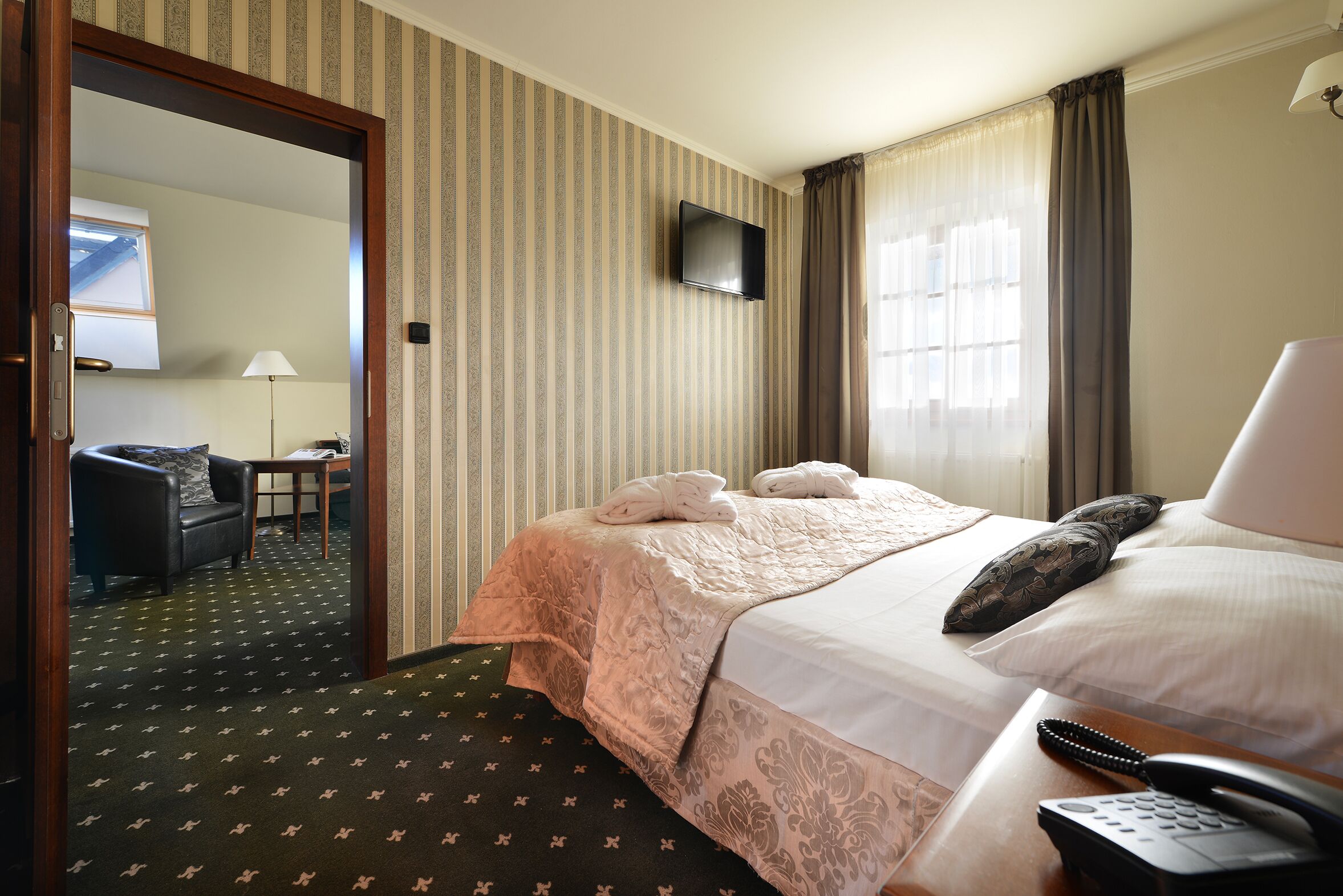 A hotel room with a bed, a bedside table with a telephone, a mirror, a window with curtains, and a view of another room.