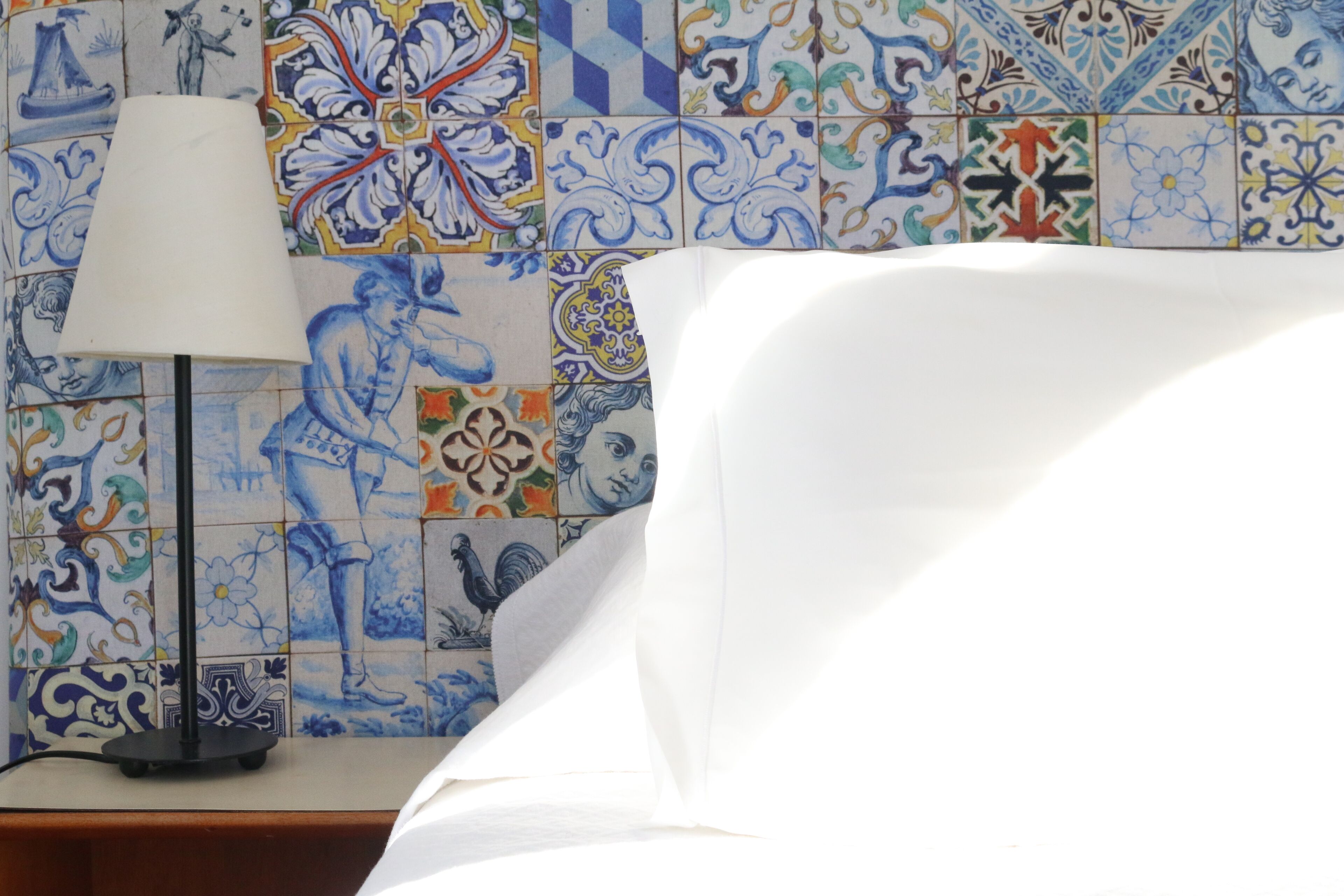 Un lit avec du linge de lit blanc, une lampe noire et un mur recouvert de carreaux aux motifs bleus et blancs.