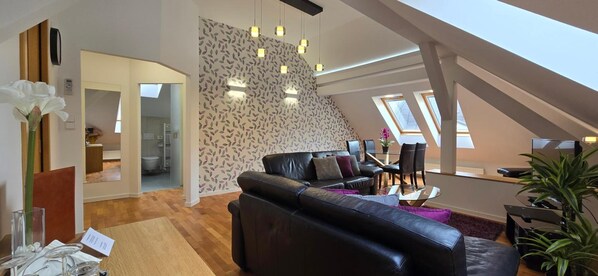 Uma sala de estar moderna com um sofá de couro preto, uma mesa de centro de vidro e um espaço de jantar com mesa e cadeiras. Há uma escada que leva a um andar superior com claraboias.