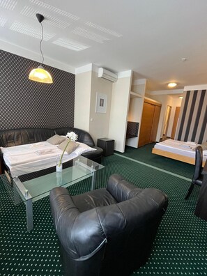 Quarto de hotel com cama, sofá, mesa de centro de vidro e uma cadeira. Há uma imagem na parede e um ar condicionado.