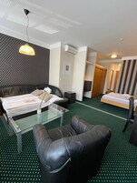 Habitación de hotel con una cama, un sofá, una mesa de centro de vidrio y una silla. Hay un cuadro en la pared y un equipo de aire acondicionado.