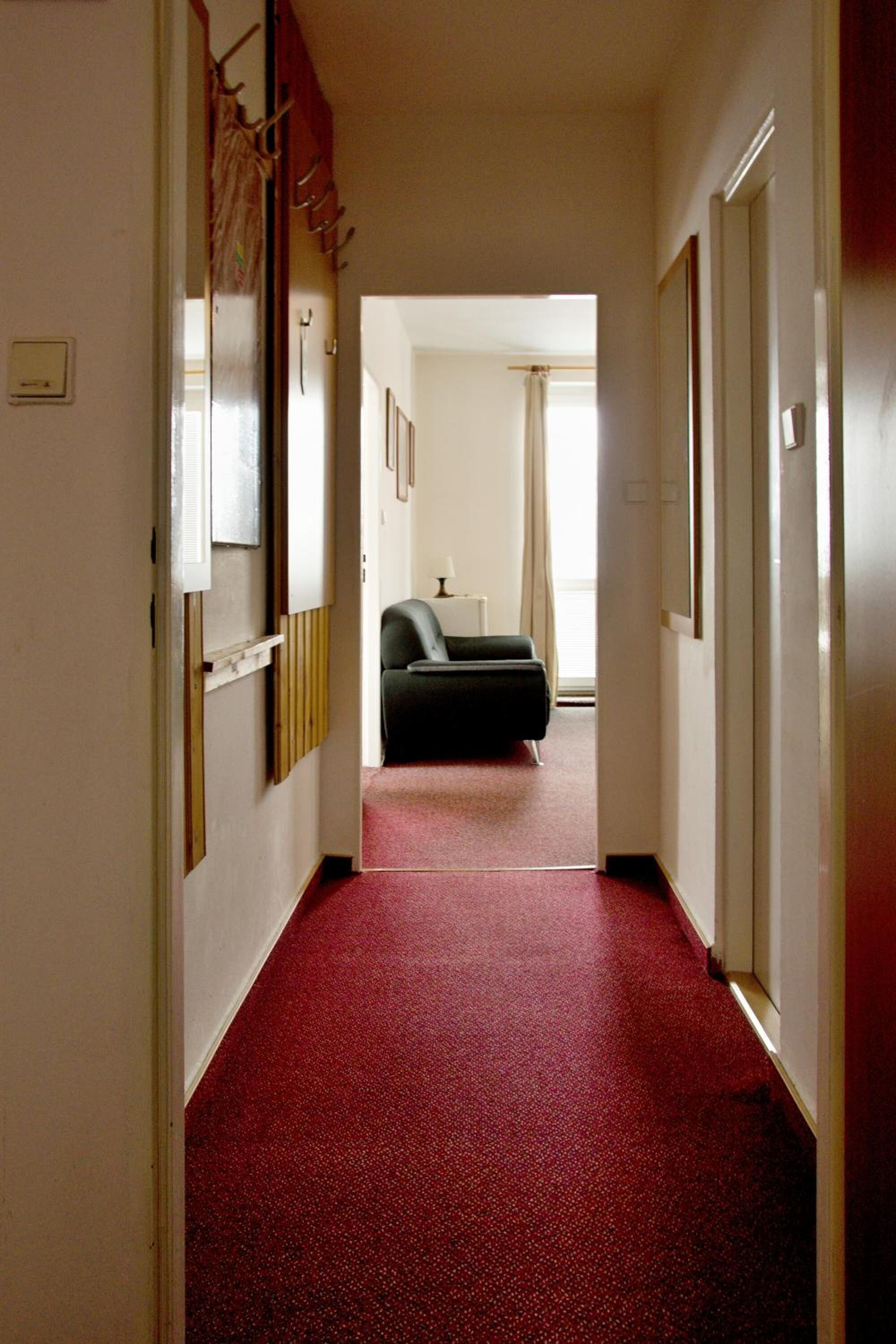 A hallway with a red carpet leading to a room with a bed and a window.