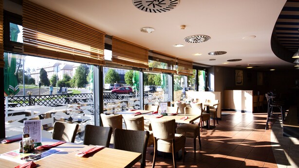 A restaurant with wooden tables and chairs, large windows with blinds, and a view of the street outside. - Hotel Arena (Liberec)